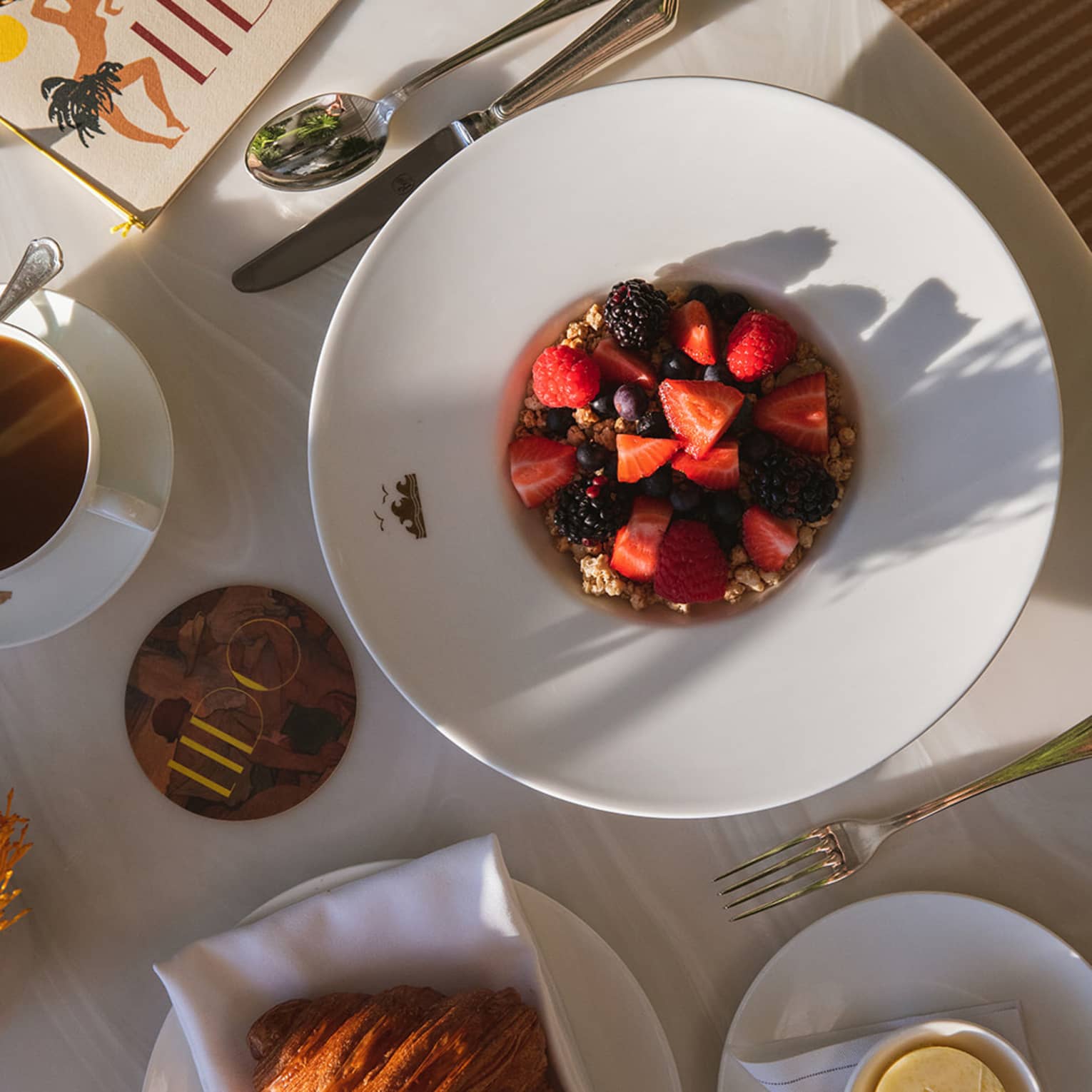 White large-rimmed bowl filled with oatmeal topped with chopped berries sites next to a crossiant, mug of coffee and other dishes on a white table