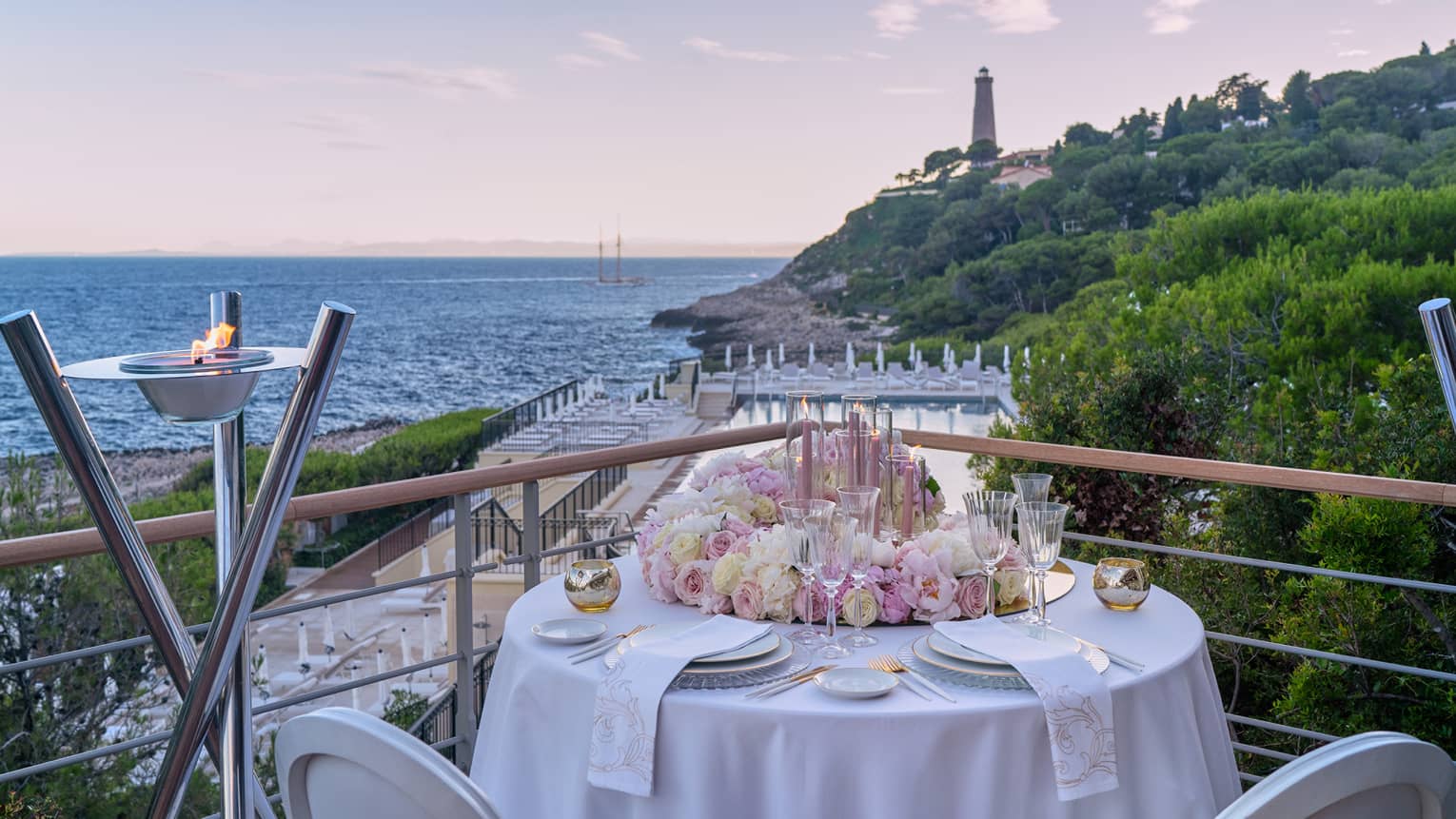 Round elegantly set white round table with pink flowers on terrace with water and lighthouse views at sunset