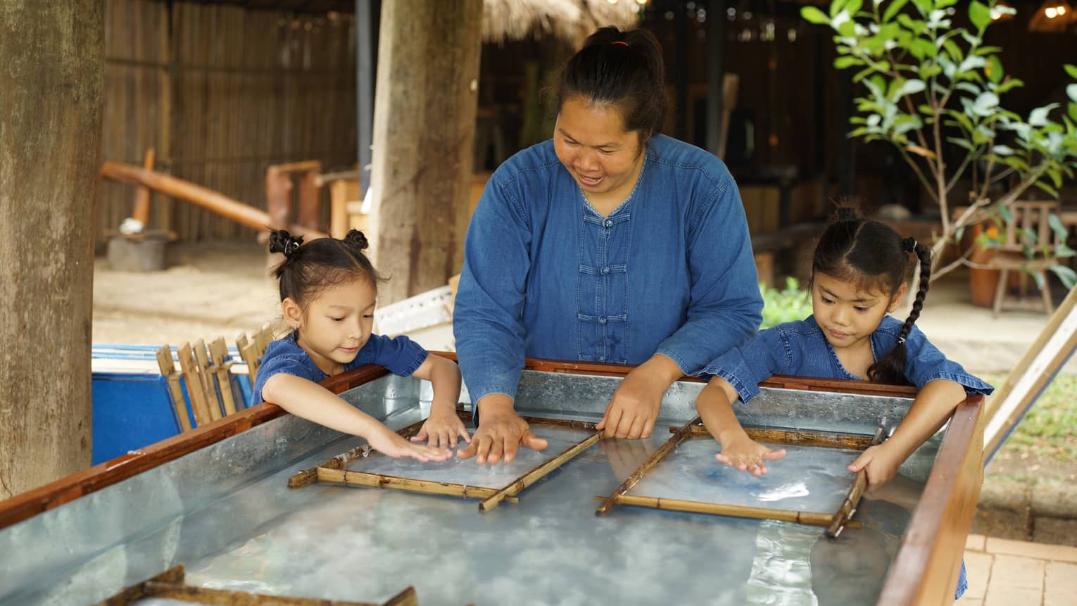 An adult stands between two children, running their hands along a clear, wet substance enclosed in two thin bamboo frames.