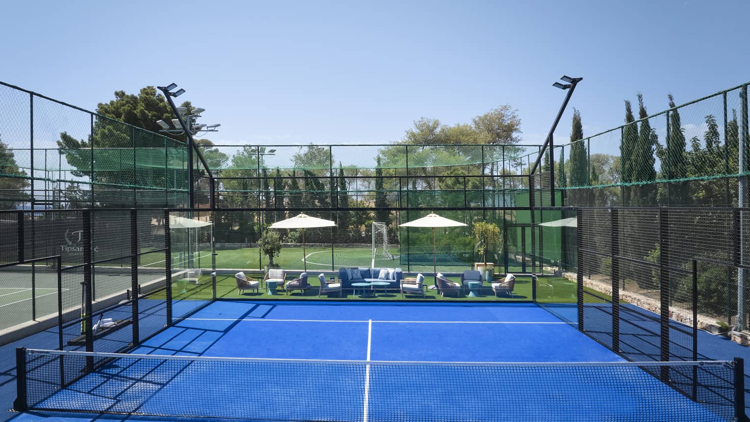 Bright blue padel court surrounded by fencing, white umbrellas and lounge chairs