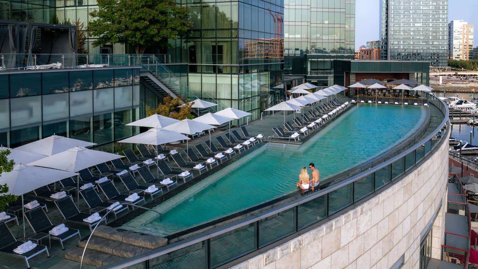 Two guests sitting on the edge of a rooftop pool overlooking the city