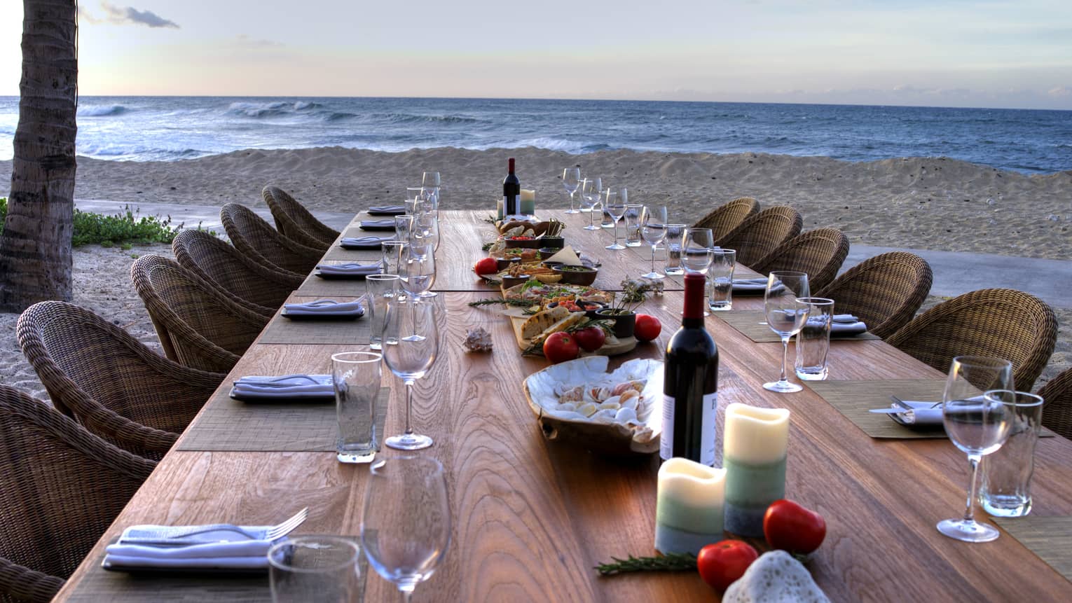 View down long wood private dining tale with wine, candles and shells, wicker chairs on beach