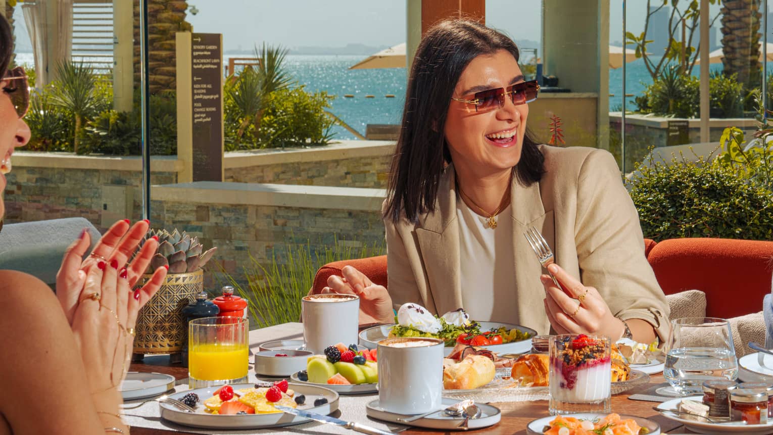 Person with shoulder-length dark hair and wearing a tan blazer, white shirt and sunglasses smiles as they sit at a table filled with food with friends on either side