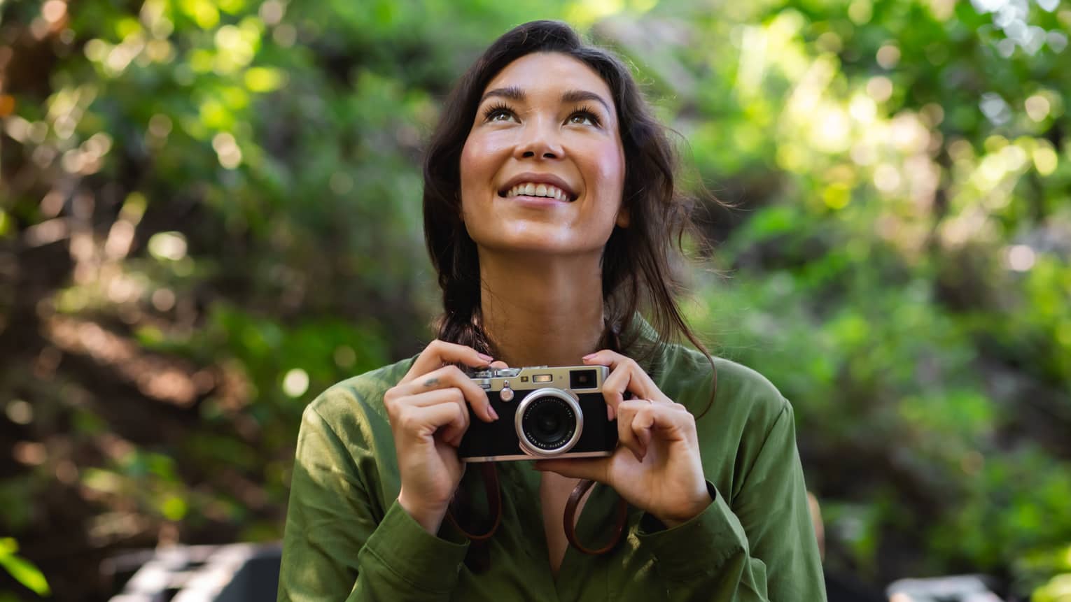 Woman with long dark hair wearing a green button-down shirt holds a camera as she looks upward in the forest