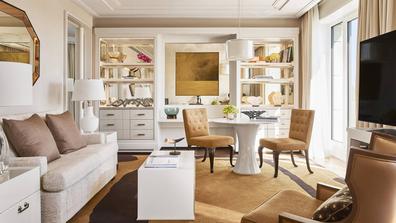Luxury hotel living room with plush seating, tan chairs, round and rectangular tables, TV, decorative shelves and large windows with beige curtains