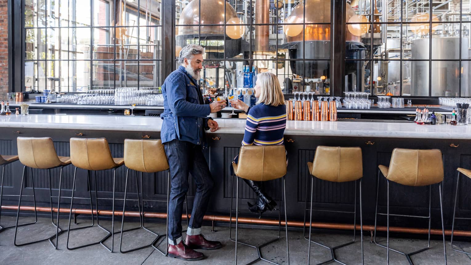 A man and woman sitting a bar with large panel windows and beige stools.