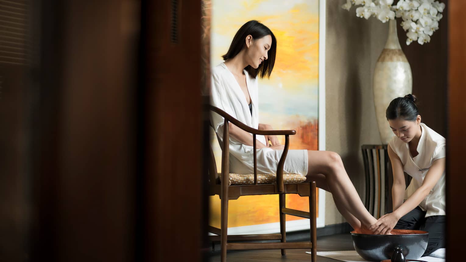 Spa staff rinses woman's feet in bowl as she sits in chair in white robe