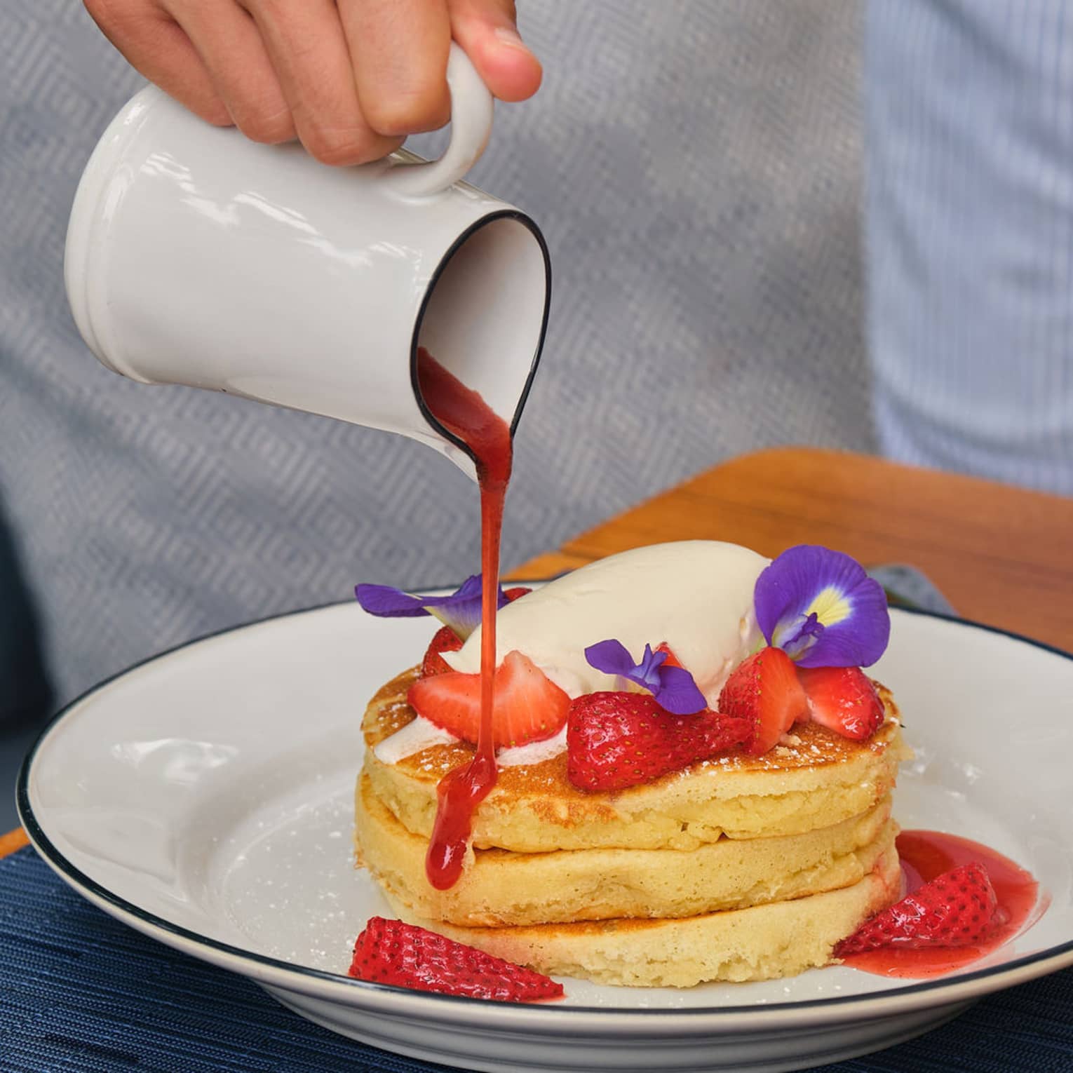 Hand ours red syrup from a white carafe onto a stack of three fluffy pancakes topped with fresh strawberries and cream and garnished with purple flowers
