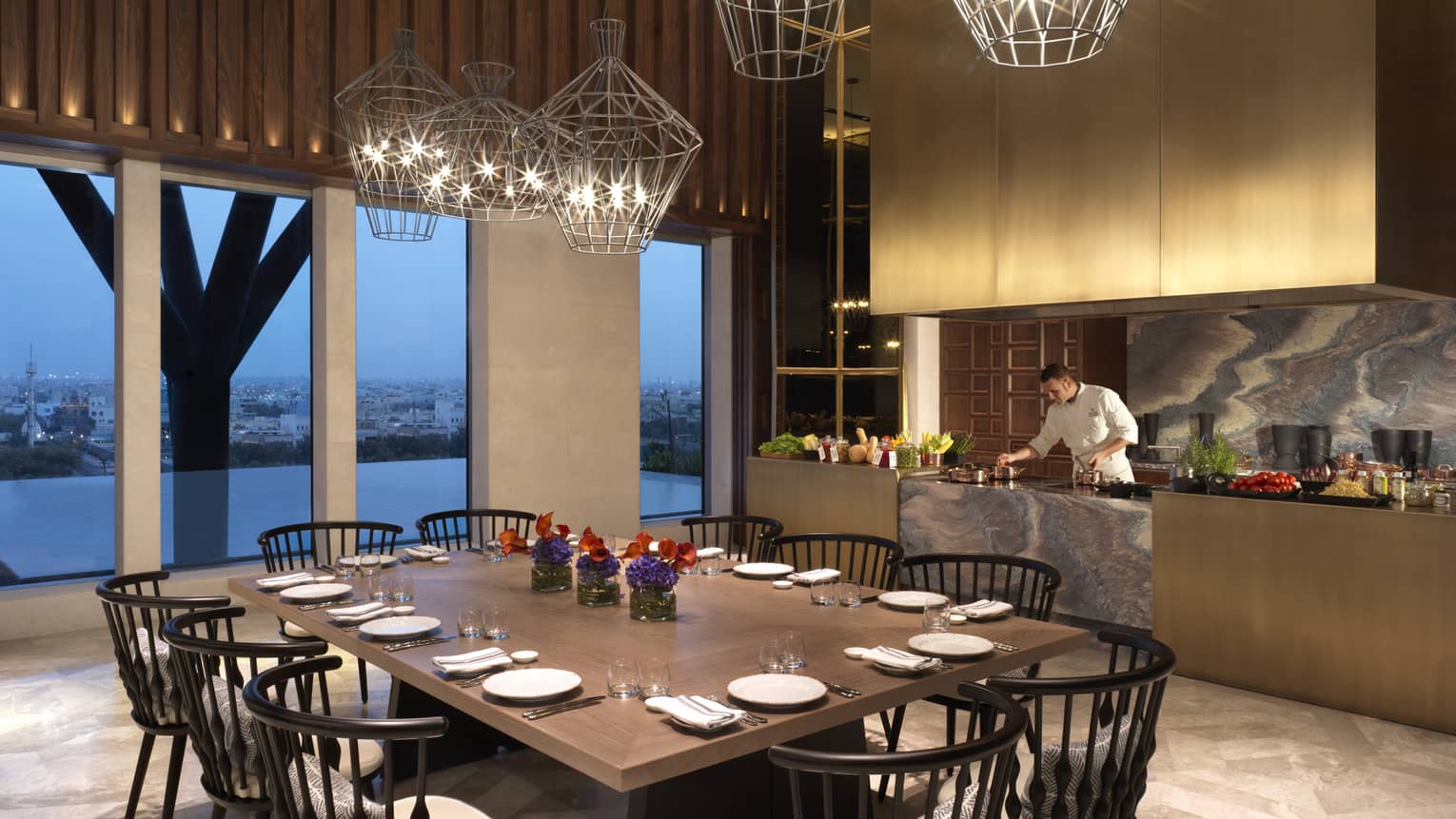 Large 10-person dining table under modern geometric wire lights, beside window and chef at marble bar