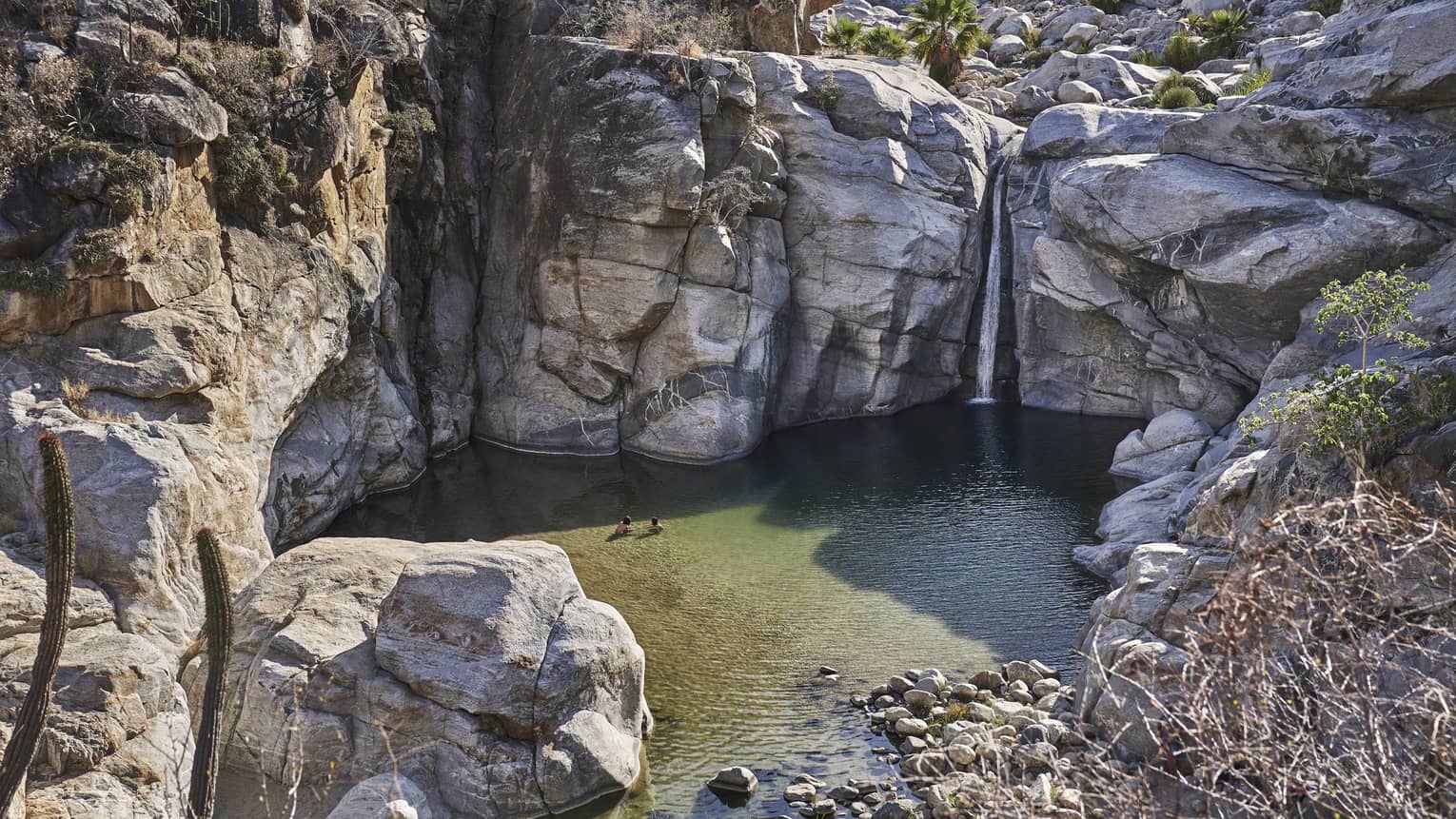 Two people wade in a small ocean lagoon surrounded by large cliffs, creating a serene and picturesque scene with clear water and rugged natural beauty.