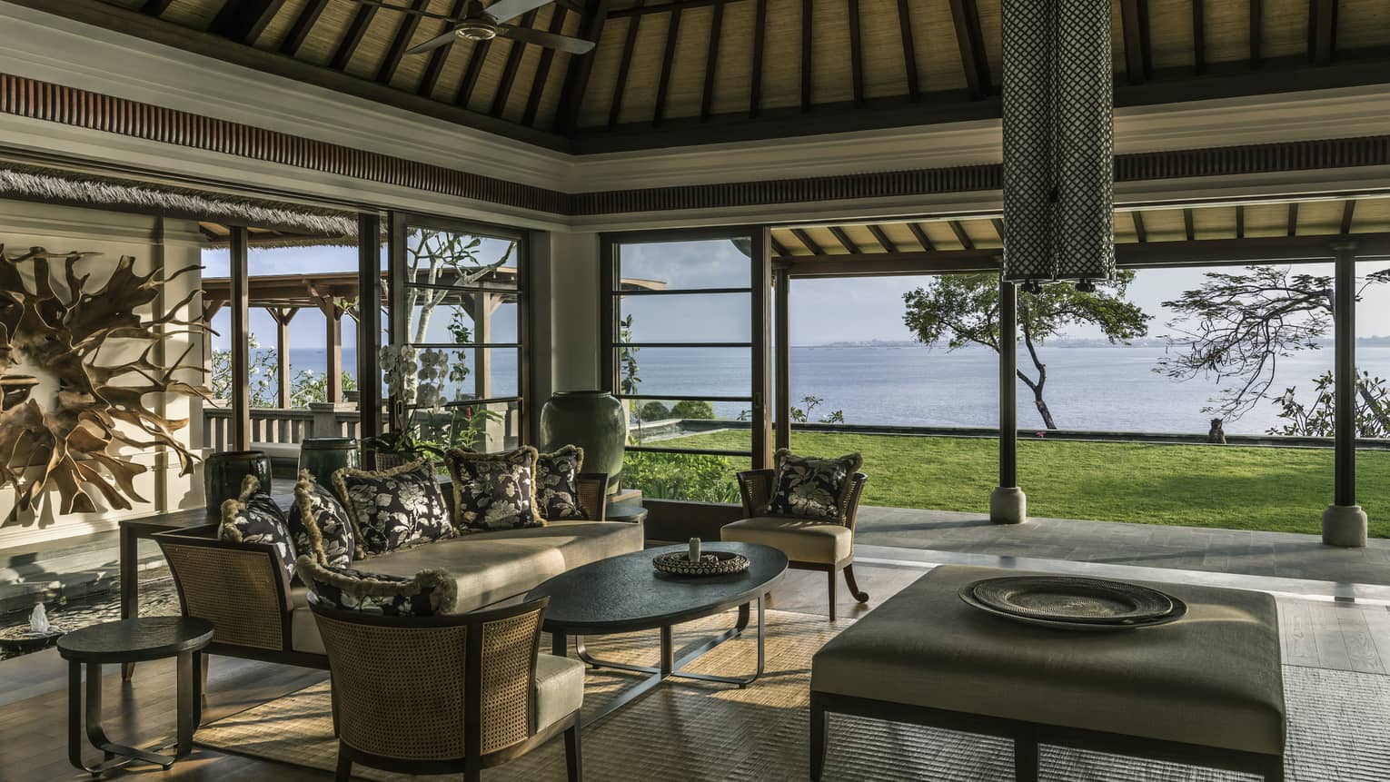 Imperial Villa open-air living room with elegant wicker sofa, chairs, large ottoman table with platter, green lawn overlooking ocean