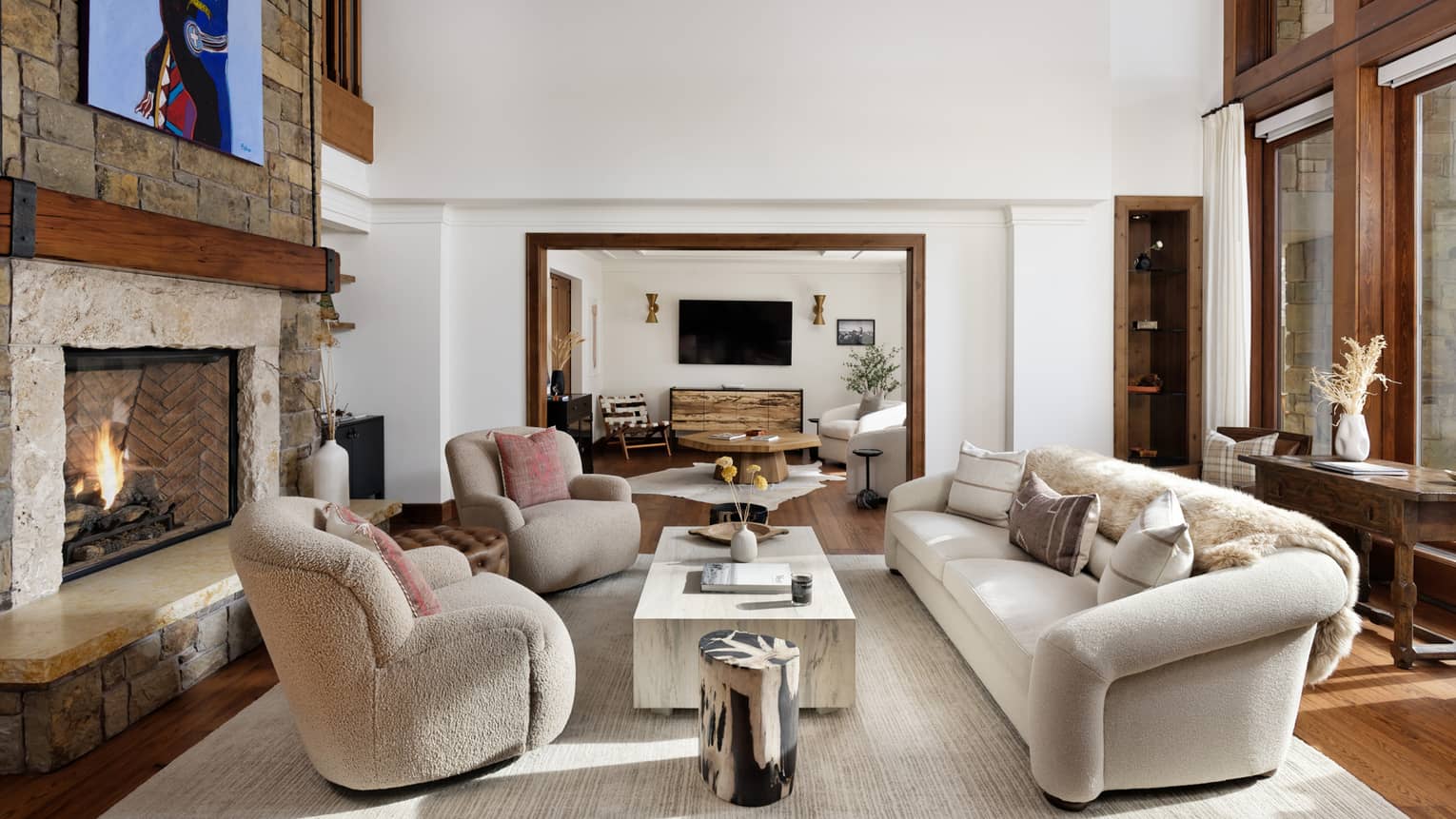 Mountain residence living room with a stone fireplace, sofa and armchairs around a coffee table, leading into a connecting seating area with a TV on the wall