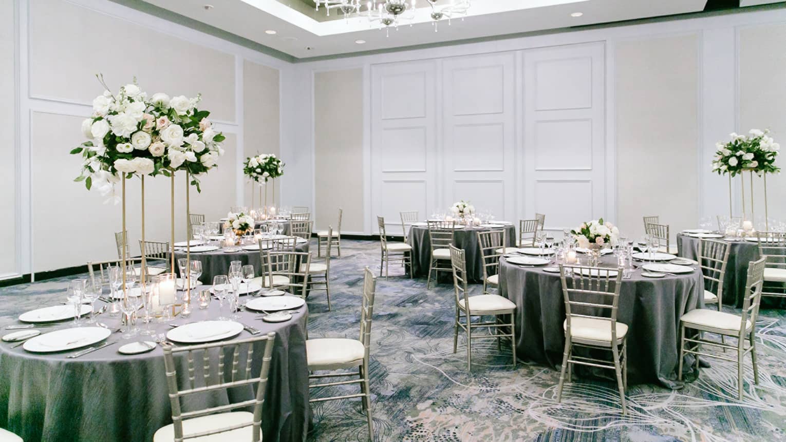 A reception area with grey clothes round tables and white wooden chairs, flower centerpieces, and a large chandalier.