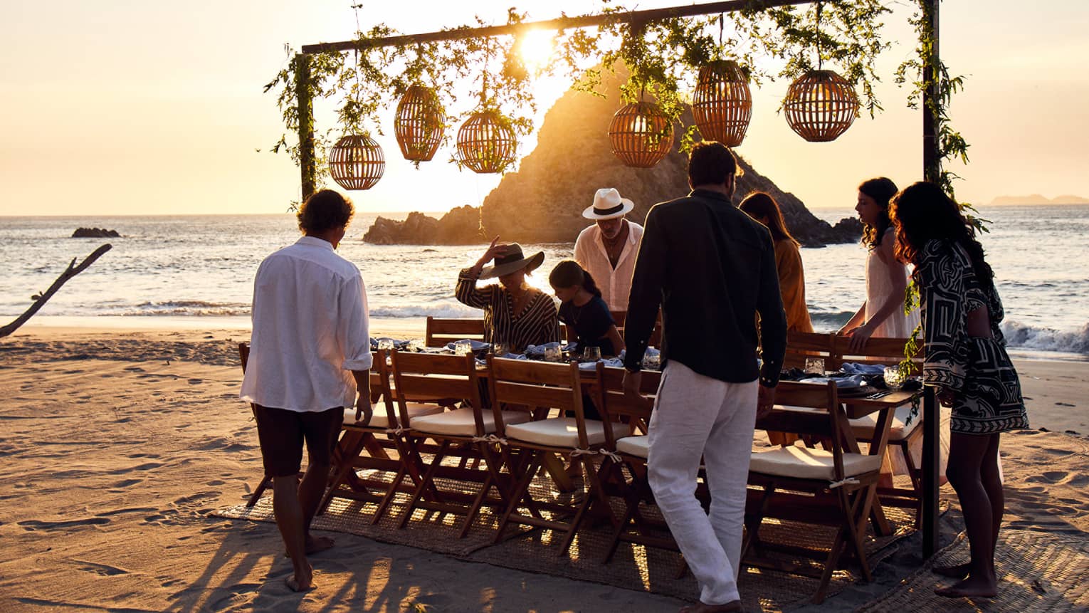 A group settles into cushioned chairs at a beachside table at dusk, lanterns and vines hanging from an attached beam above.