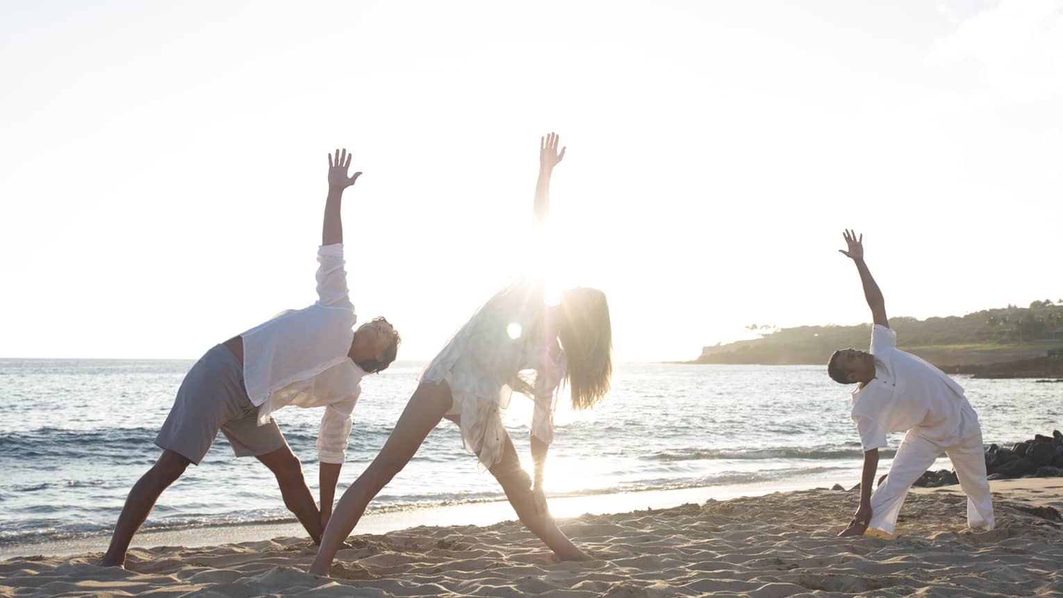 Two people in white beach shirts and shorts do a yoga pose with yogi at sunrise on beach