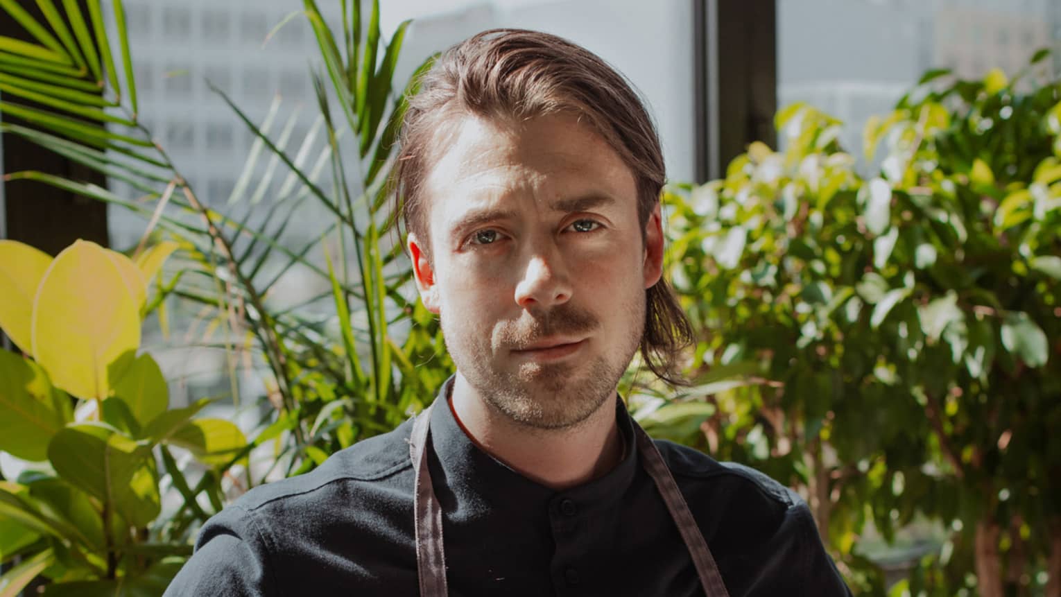 Executive Chef Jason Morris standing in front of window and plants in black chef?s coat and dark grey apron