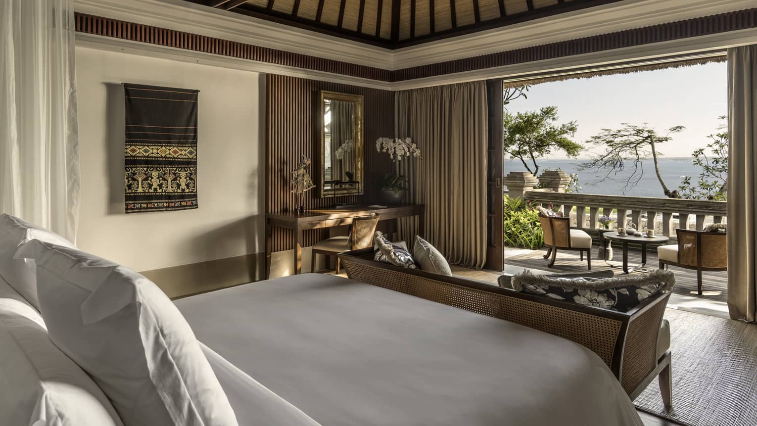 Premier Ocean Villa bedroom with bed and wicker bench facing curtains, open sliding doors to patio