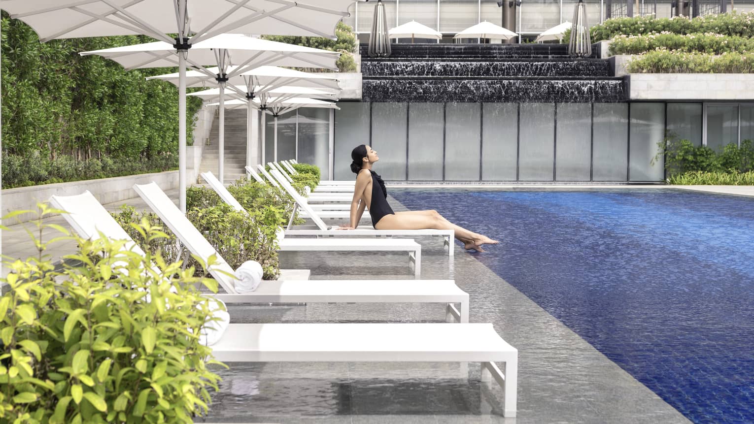 A woman in a black bathing suit relaxes on a chaise by the pool