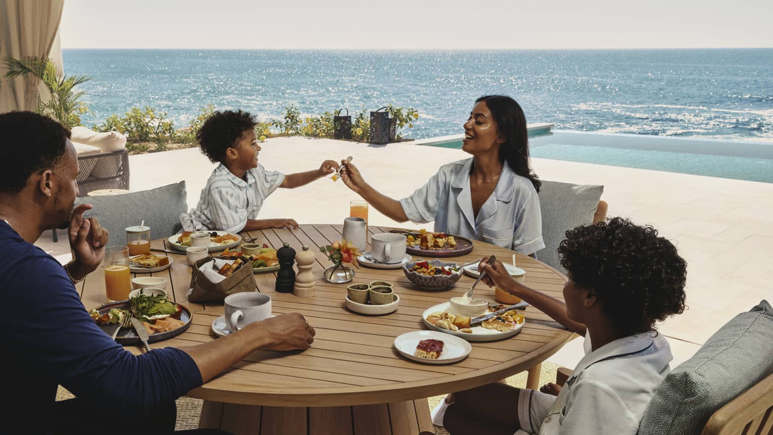 Man, woman and two young boys eat at wooden table on ocean-view terrace at luxury resort