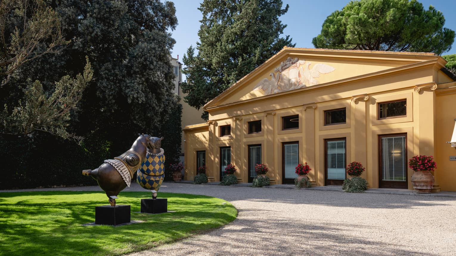 A small building with large windows on manicured grounds complete with potted plants, a drive and sculptures, with large trees in the background