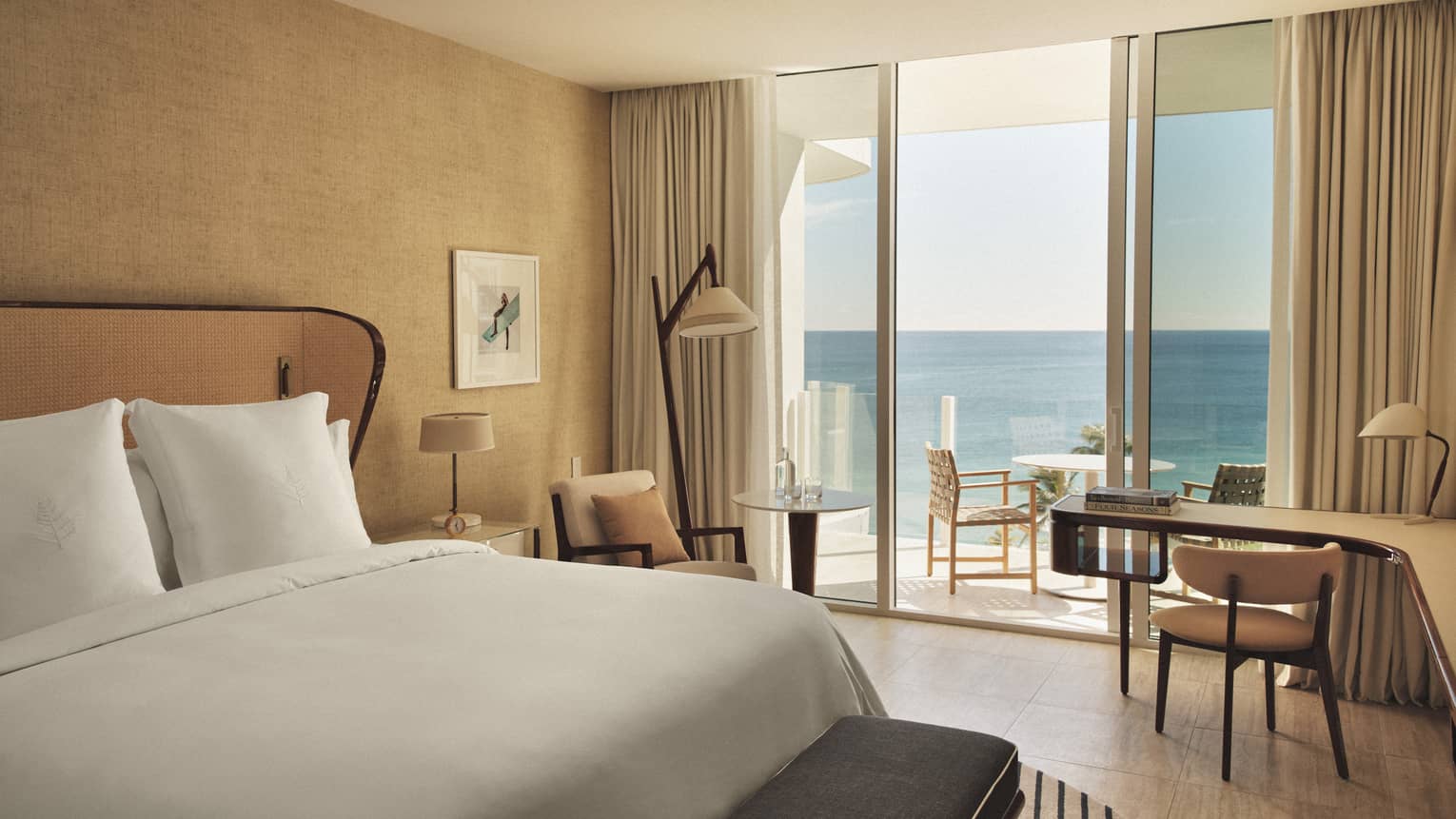 A hotel room with sliding glass door, bed with white sheets, long curved desk, two lamps, two chairs and outdoor seating.