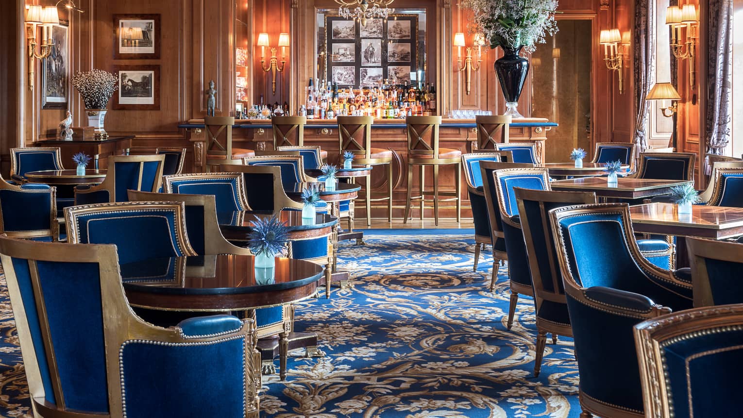 Le Bar des Bergues lounge with elegant blue velvet chairs with wood trims around cocktail tables, crystal chandelier, bar