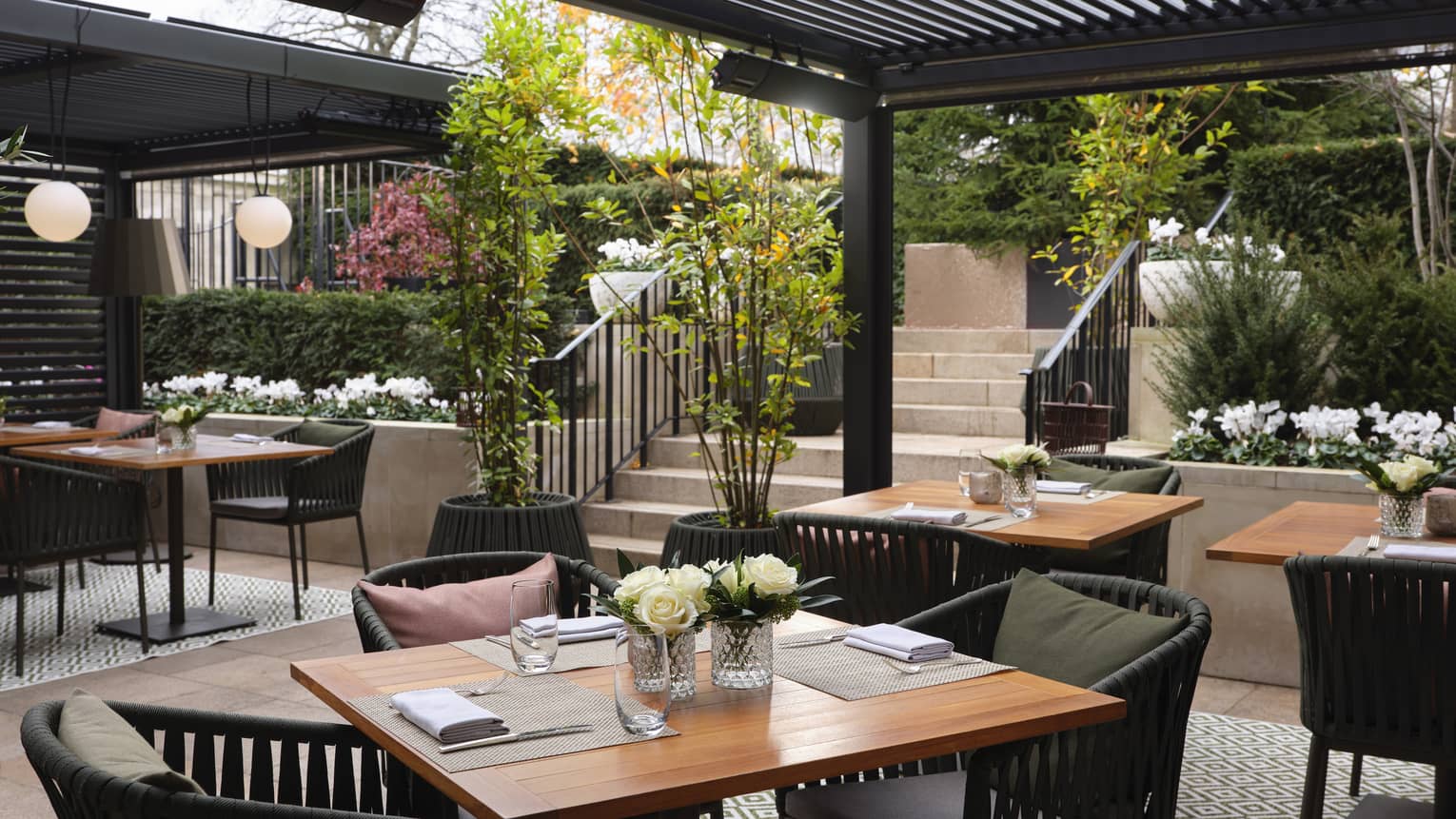 Set square wooden tables, dark rattan chairs, outdoor diamond-patterned rug under pergola