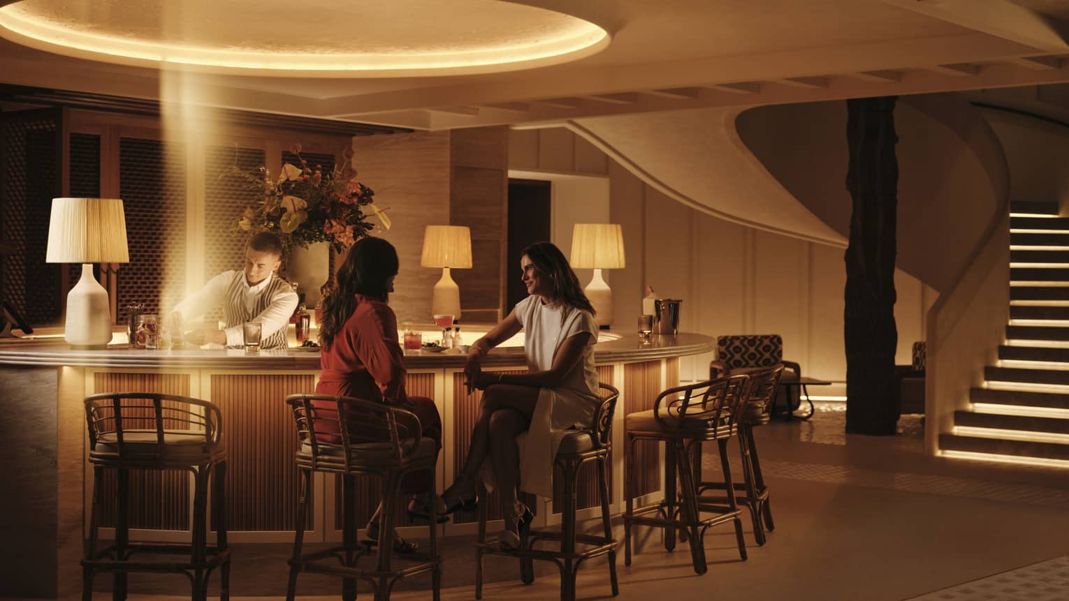 Two smiling guests sit at a warmly lit circular bar at night as a bartender prepares cocktails in a stream of golden light.