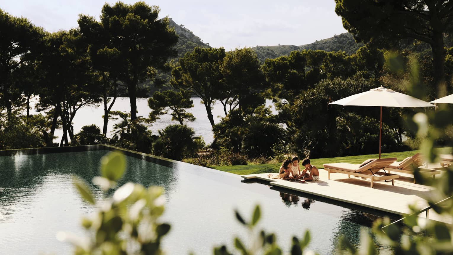 young children play on pool deck next to large resort pool, surrounded by push pine trees