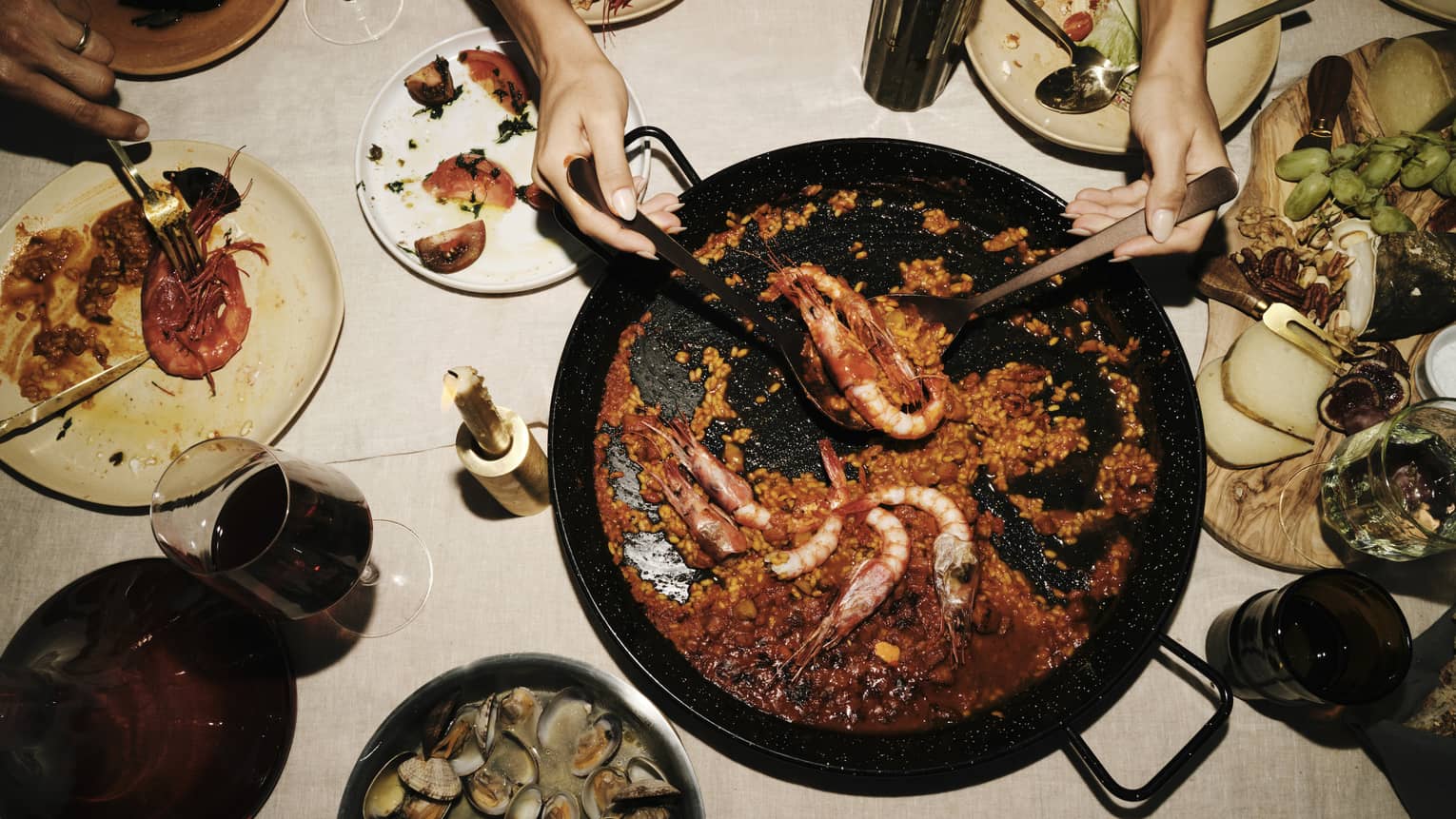 Two hands use large spoons to lift prawns from a saucy paella dish set amid plates of seafood, cheese and fruit.