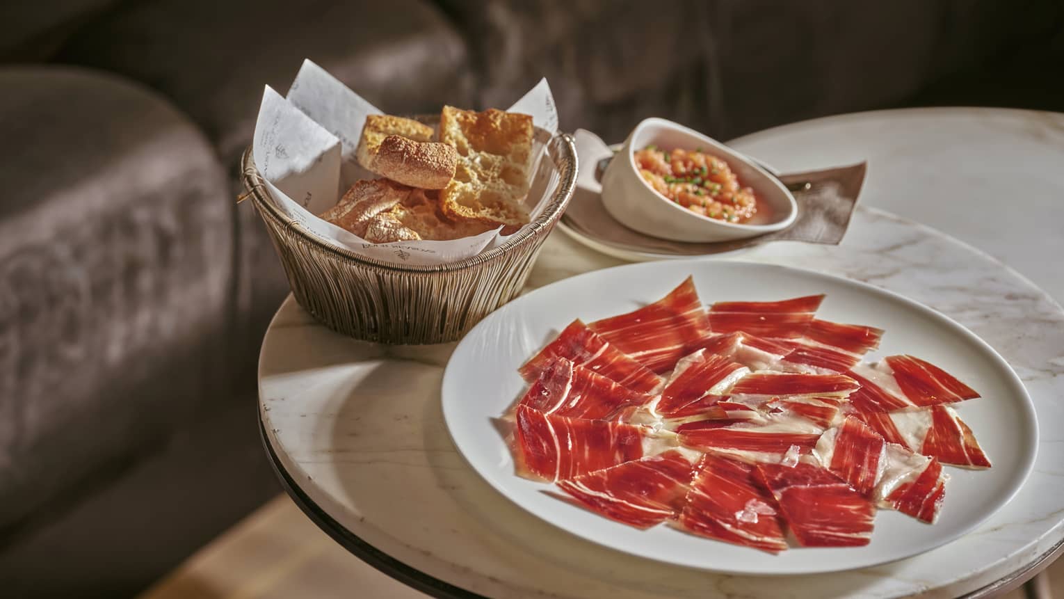 Plate of Iberico ham slices beside bowl of crackers on small marble-top table