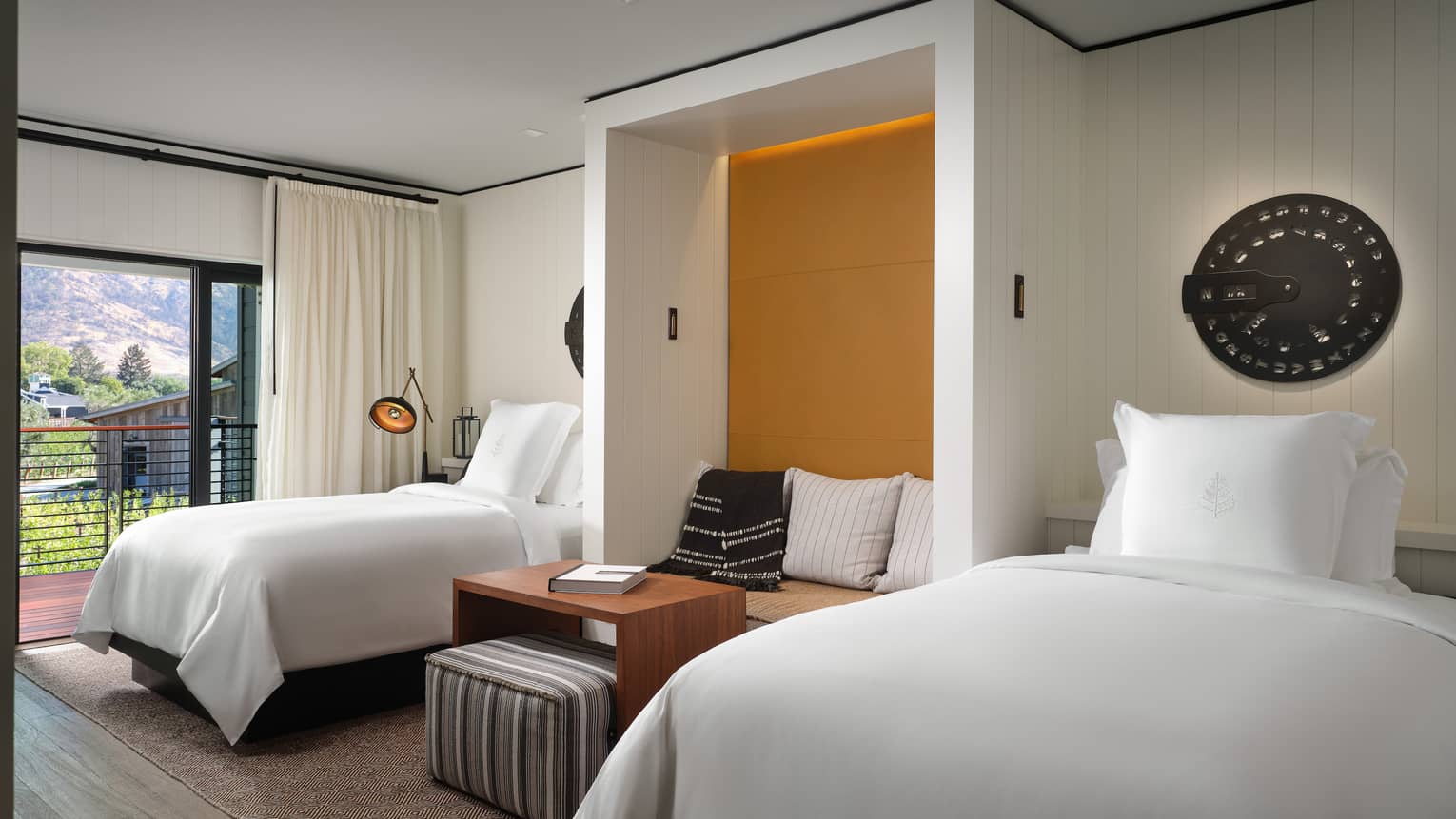 Luxury resort room with two beds, built-in seating nook with mustard accent wall, striped ottoman, decorative wall art and mountain-view balcony 