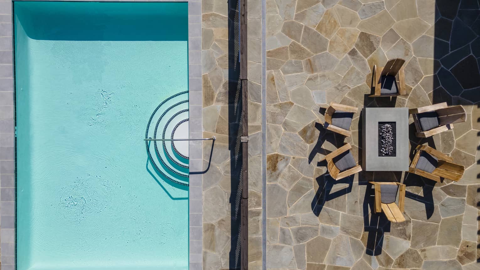 Aerial view of a luxury resort villa backyard with a rectangular pool, stone patio and fire pit surrounded by wooden lounge chairs