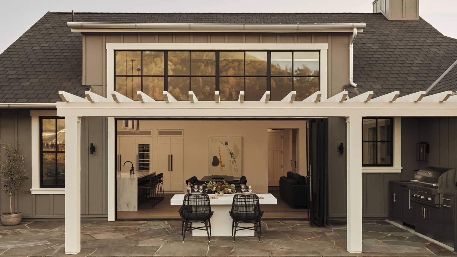 Outdoor dining area of a residential villa with pergola, white table and black wicker chairs, opening to a kitchen and living room through large folding doors