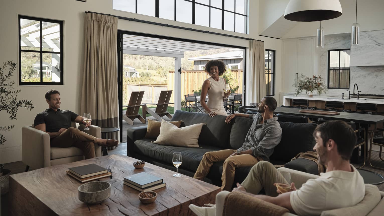 Friends gather around a table in a living room, seated on a couch and club chairs, with a view of the villa's outdoor area.
