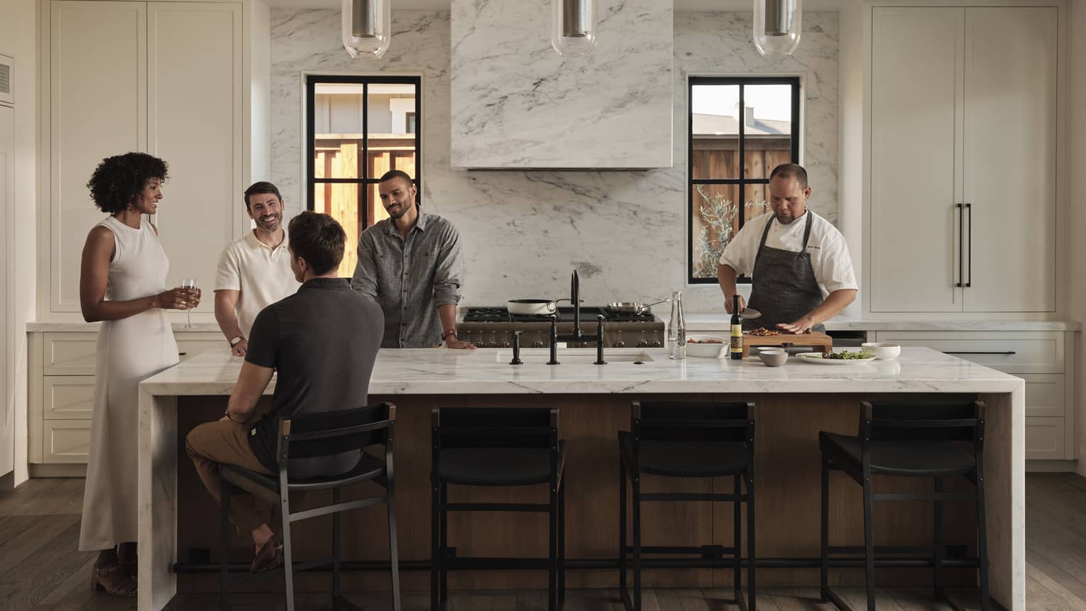 A group of guests gather at one end of a long kitchen island while a chef chops ingredients at the other end of the island.