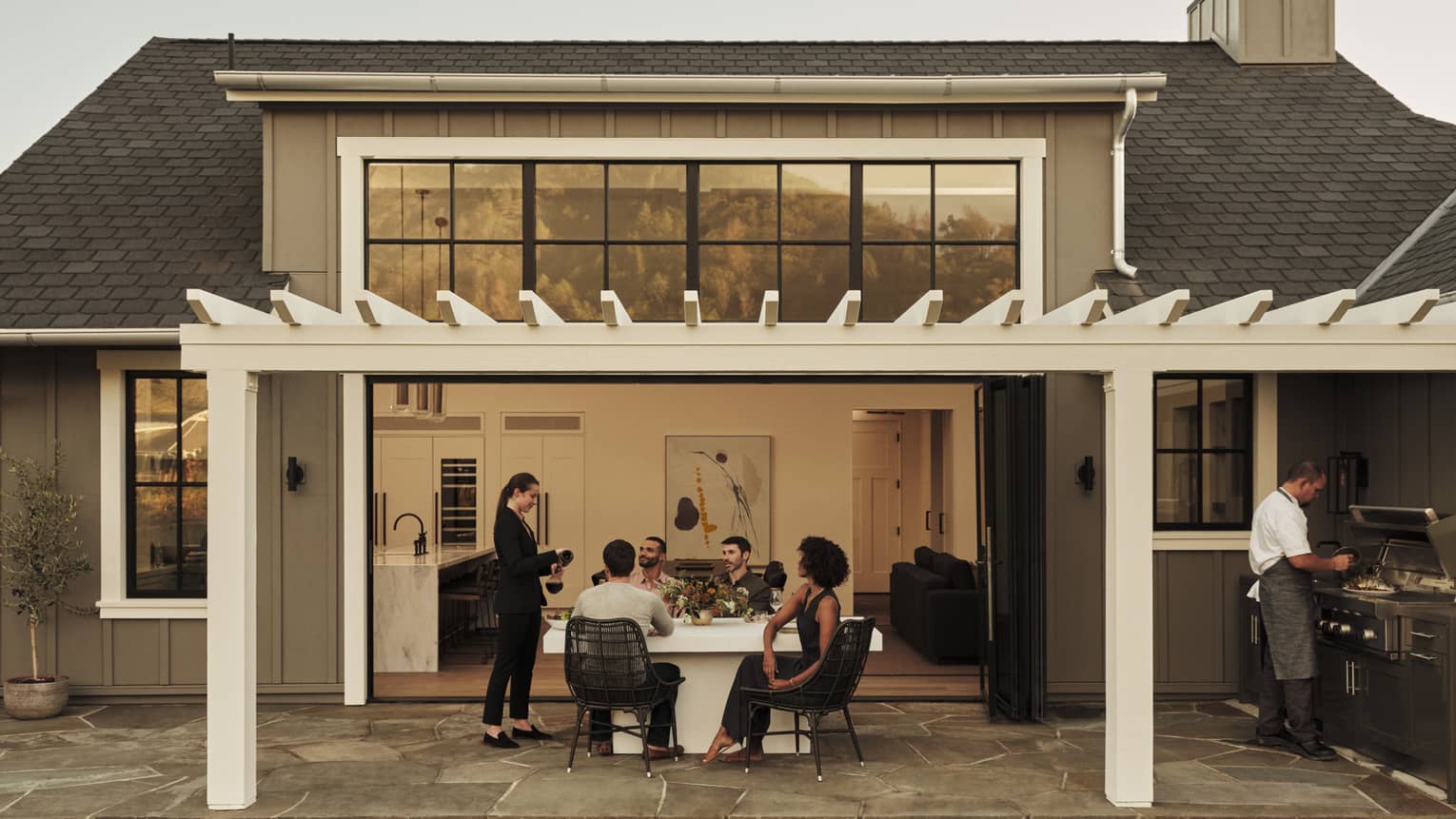 Four guests sit at an outdoor table and watch a server decant a bottle of wine; a chef prepares food at a barbecue nearby.