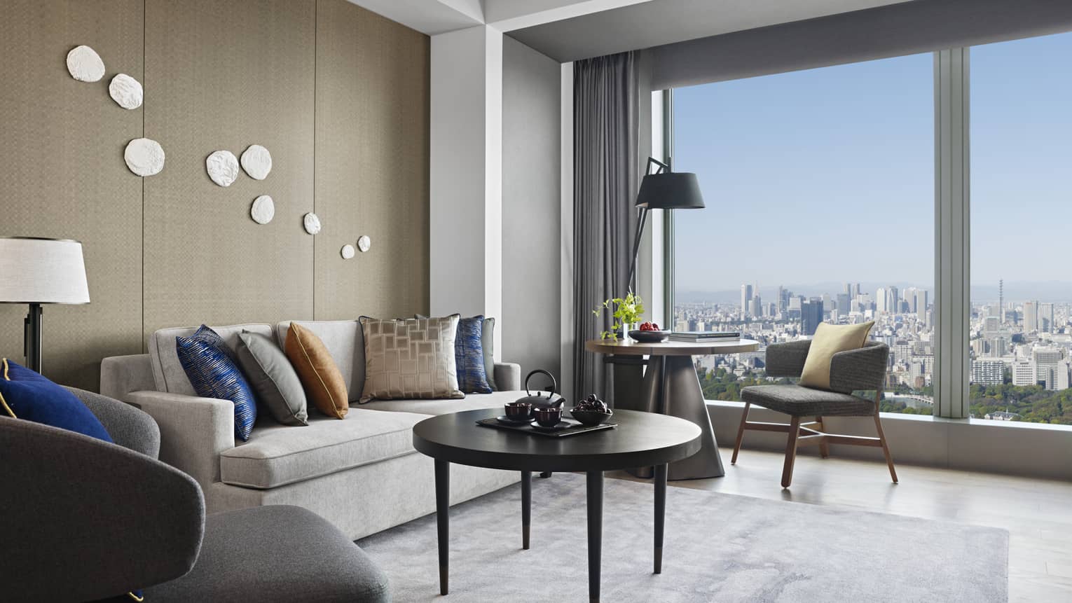 Modern living area with a grey sofa, decorative cushions and a round table, featuring floor-to-ceiling windows with expansive city views