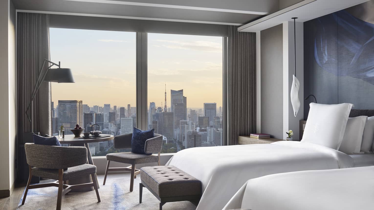 Guest room with two beds, tufted bench, seating area and floor-to-ceiling windows offering panoramic city views