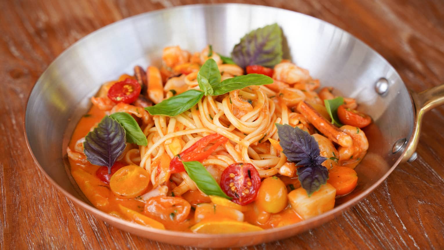 A metal skillet contains long noodles topped with colorful ingredients and garnishes.