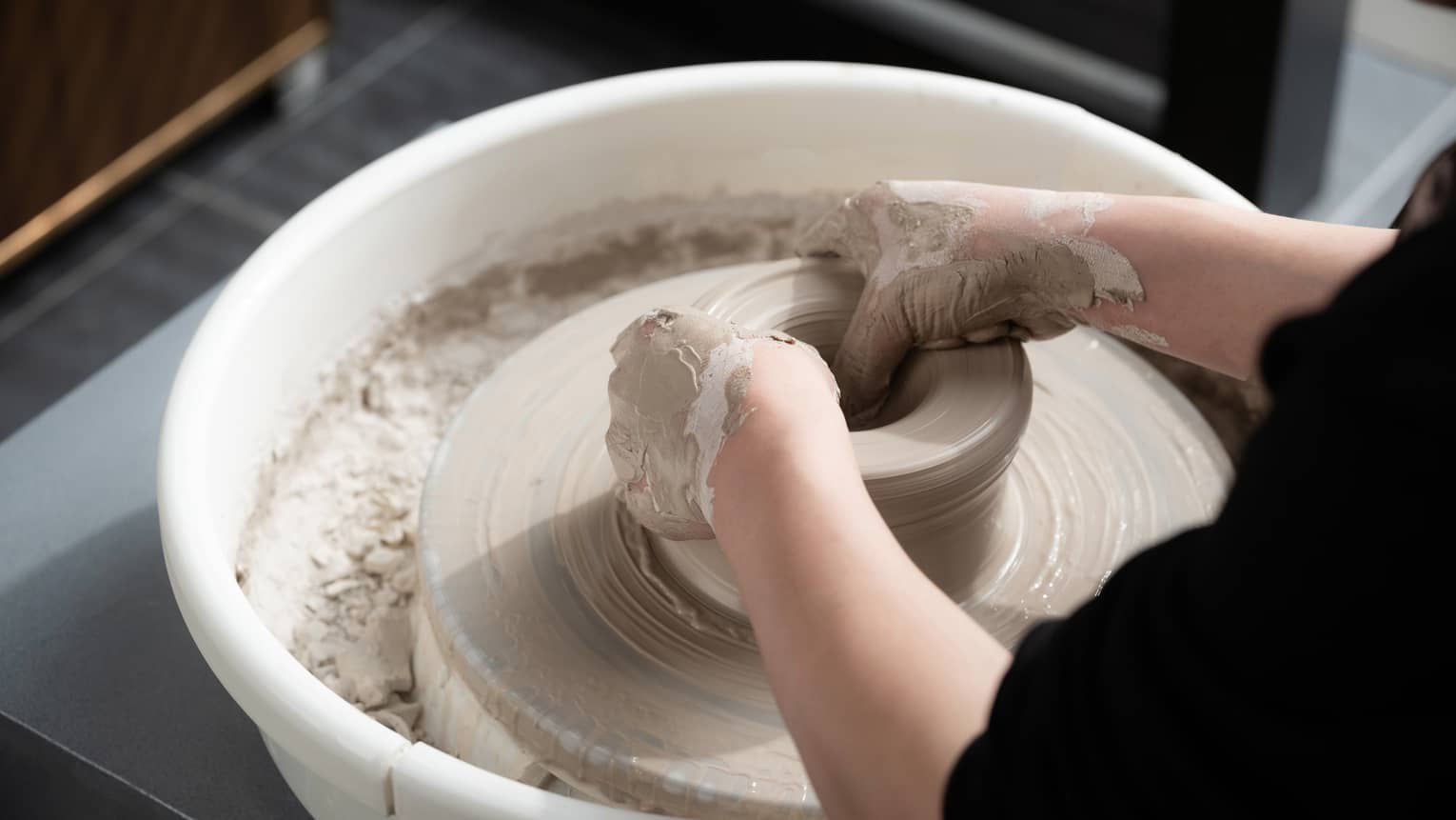 Close-up of two hands covered in wet beige clay shaping a pot on a spinning pottery wheel. The potter is wearing black.
