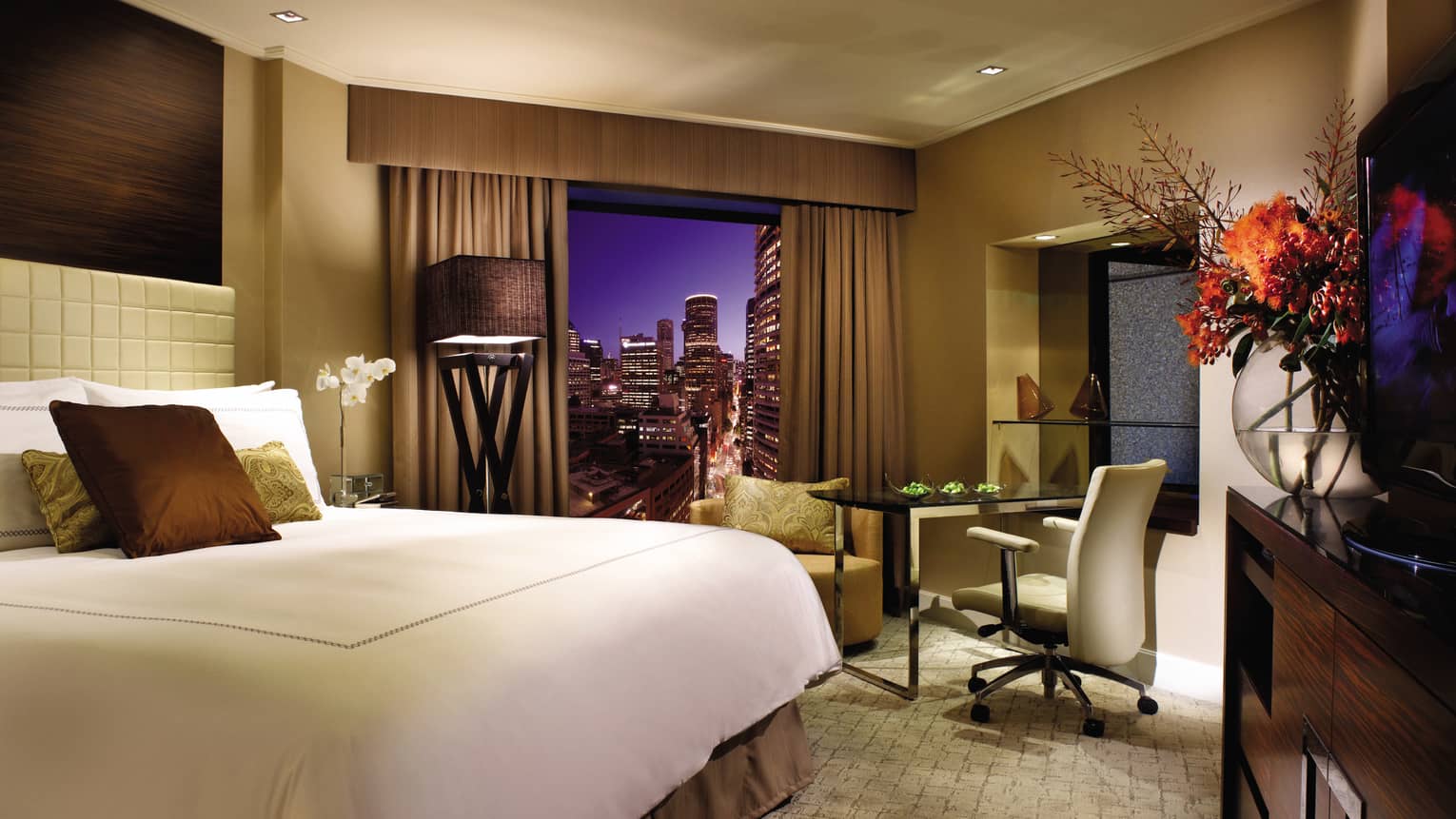 Deluxe City Room at night, bed with brown silk pillows, large glass vase with flowers, window with city view