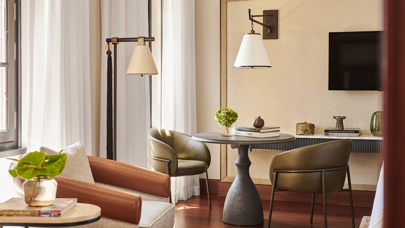 Corner of a hotel room, complete with a round table and two leather chairs, two lamps, an armchair, TV and windows