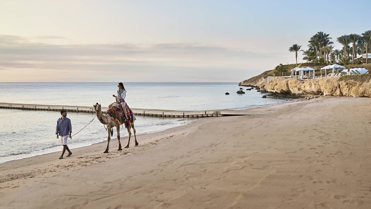 A woman rides a camel, led by a man, along the beach at Four Seasons Sharm El Sheikh