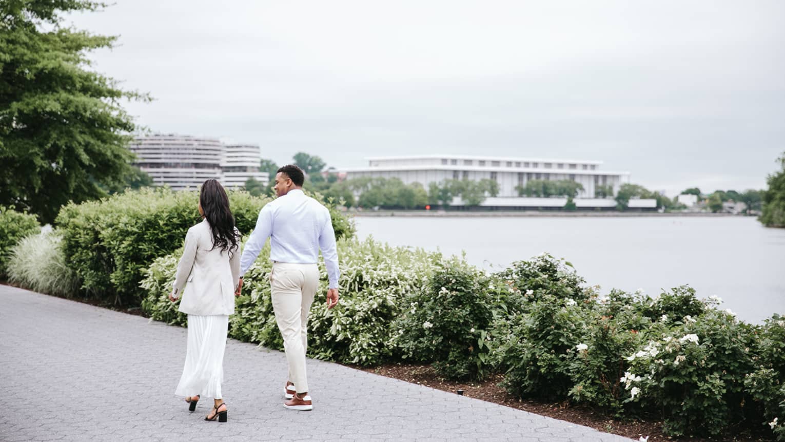 Rear view of a casually dressed couple walking along a shrub-lined brick path by the water under a bright-white sky.