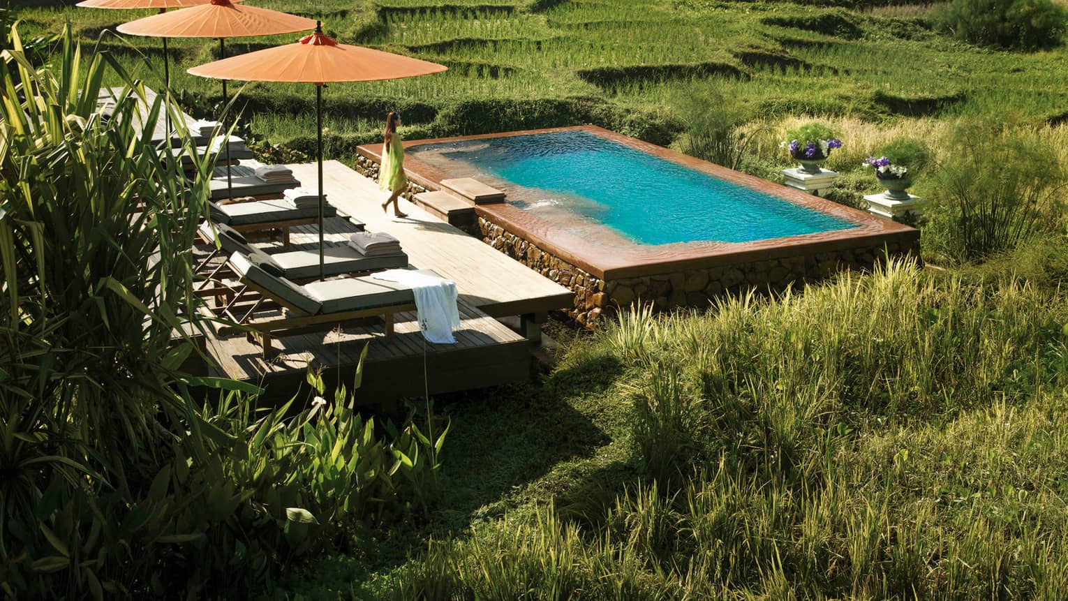 Woman in flowing dress walks past three patio umbrellas to infinity pool in rice plantation