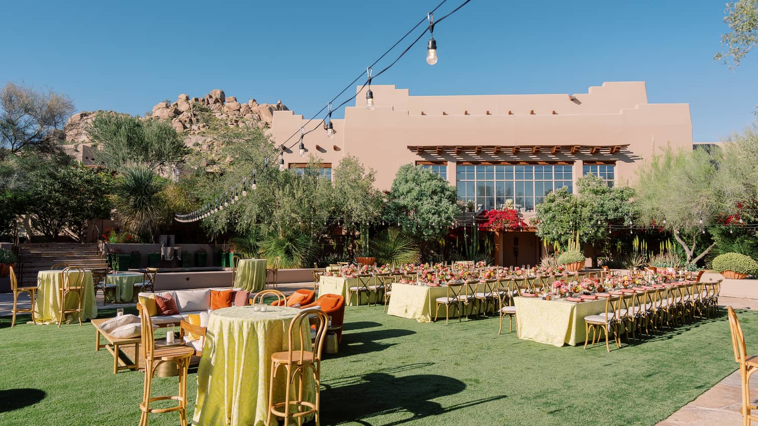 An outdoor venue with tables and chairs with place settings and a string of lights above