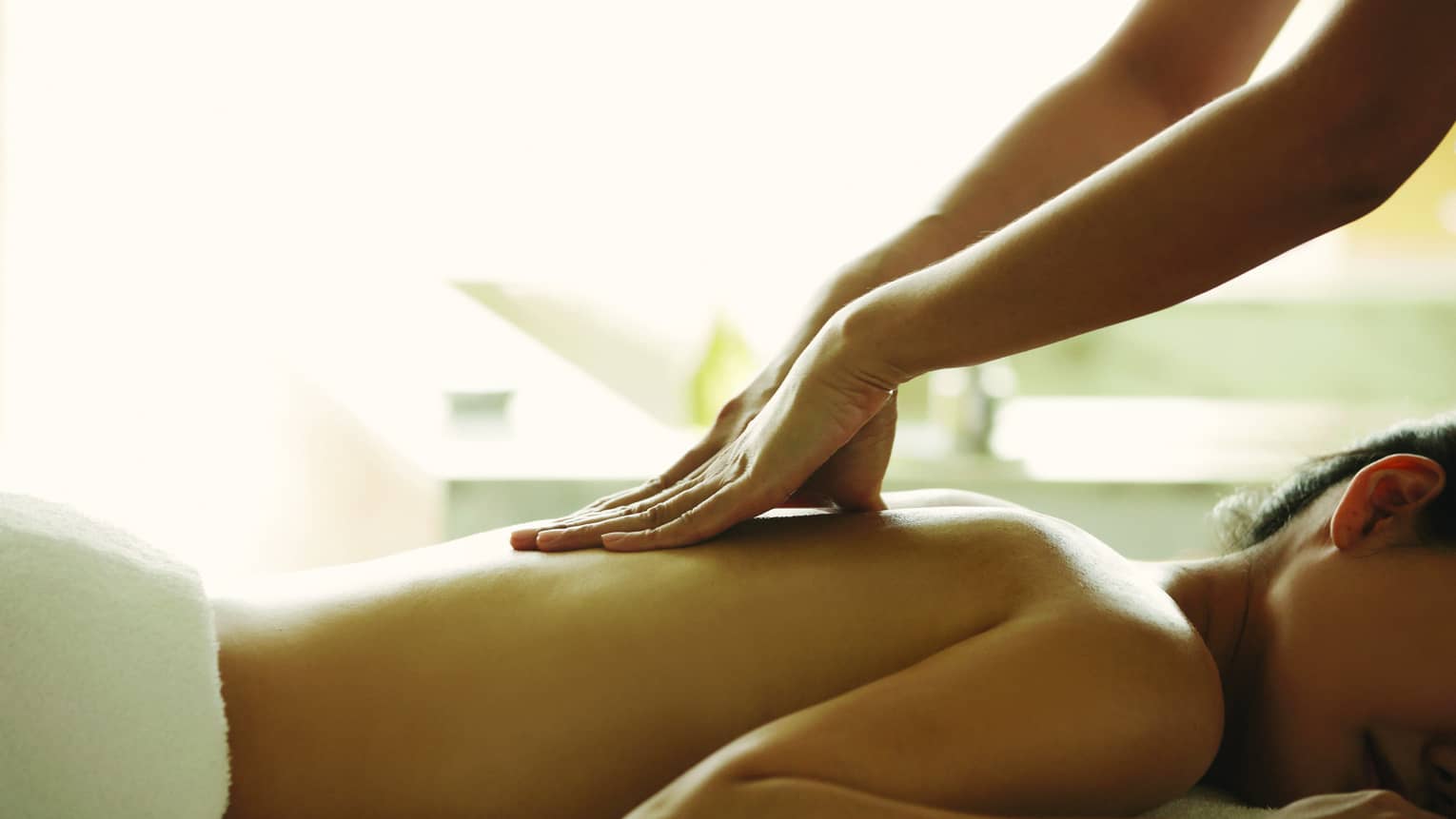 Masseuse reaches over, massages woman's bare back as she lies on spa table