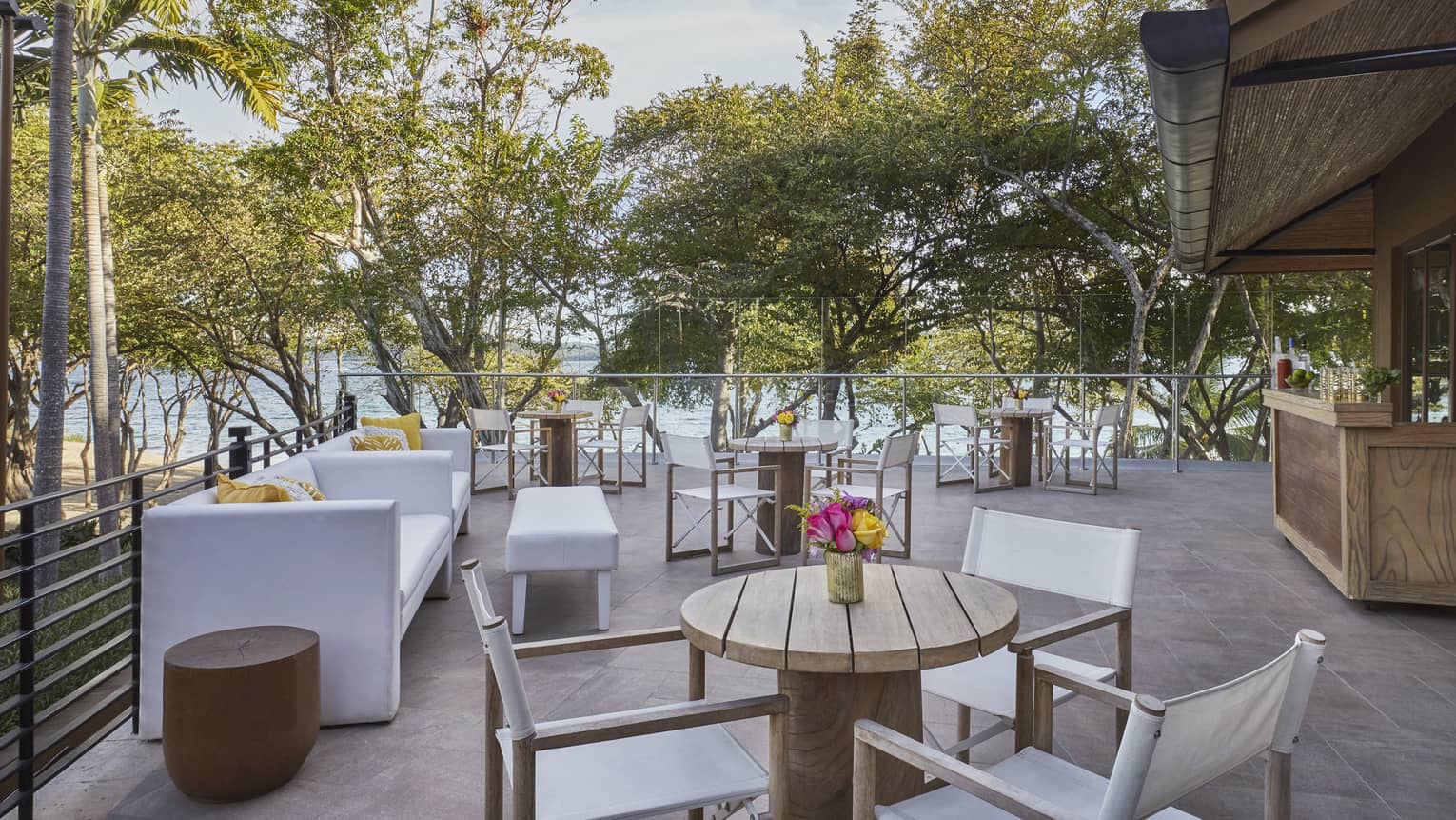 Outdoor terrace overlooking the beach with white sofas, various wooden tables and white chairs and an outdoor wooden bar