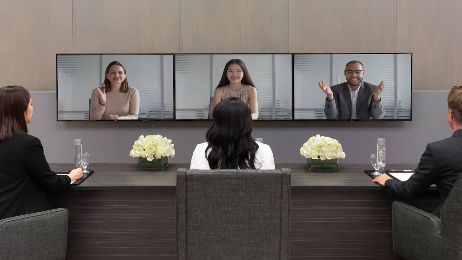 Several people in business clothes sit in front of a series of screens in a virtual meeting.
